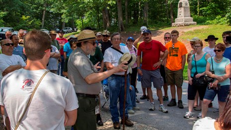 A park ranger presents a program on the battlefield. He is surrounded by dozens of visitors.