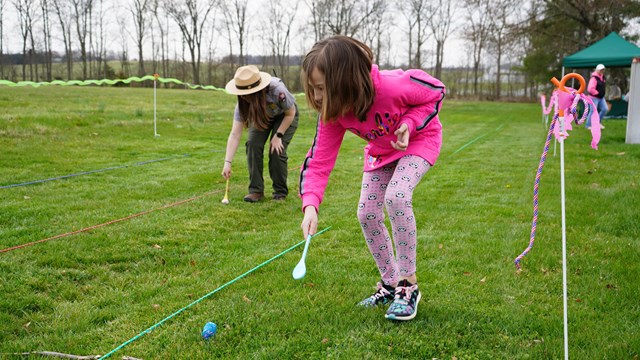 A child and a park ranger roll wooden eggs along a grassy path.