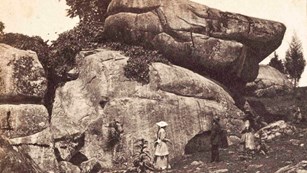 A black and white photo of two women and one man in front of a very large stack of boulders.