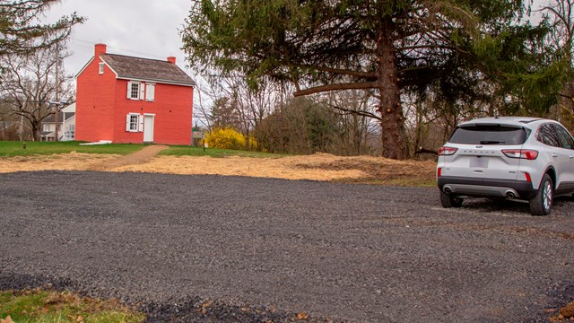 A dark gravel parking lot & white SUV are in front of a large pine tree & red brick house.