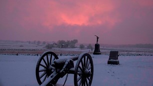 Snow covers the battlefield, a cannon and two monuments are in the center and pink and purple clouds