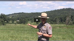 A park ranger points across a green field to two hills in the distance.
