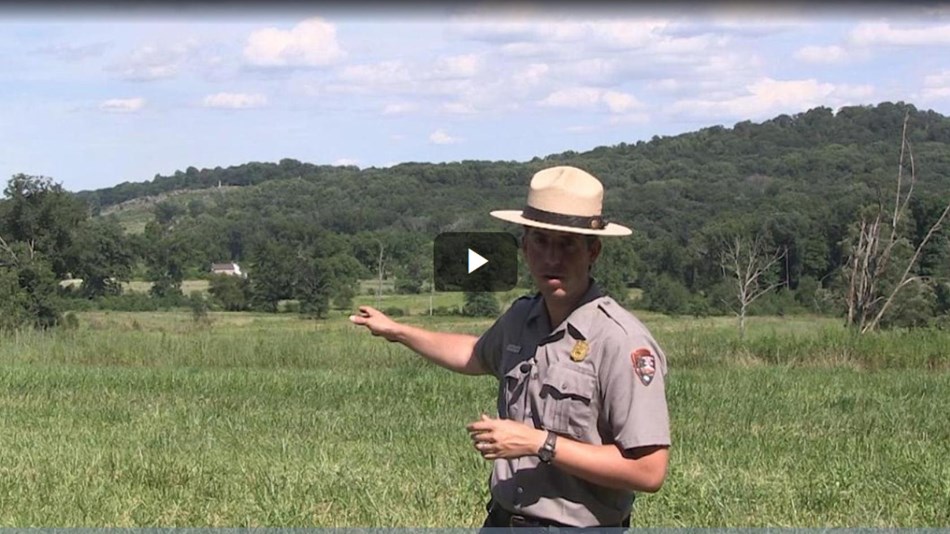 A park ranger points across a green field to two hills in the distance.