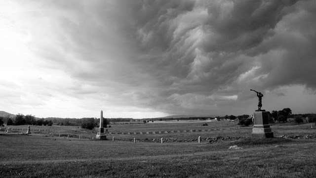 A black and white photo of storm clouds over the battlefield, two monuments are in the foreground.