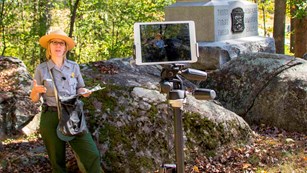 A park ranger stands beside a monument as she presents a virtual program via a digital tablet.