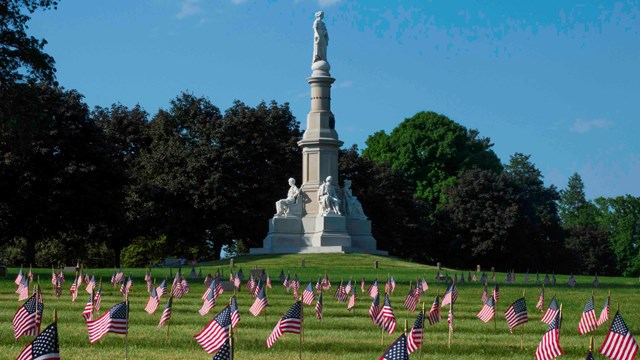 A white granite statue stands in the center of a cemetery decorated with small United States flags.