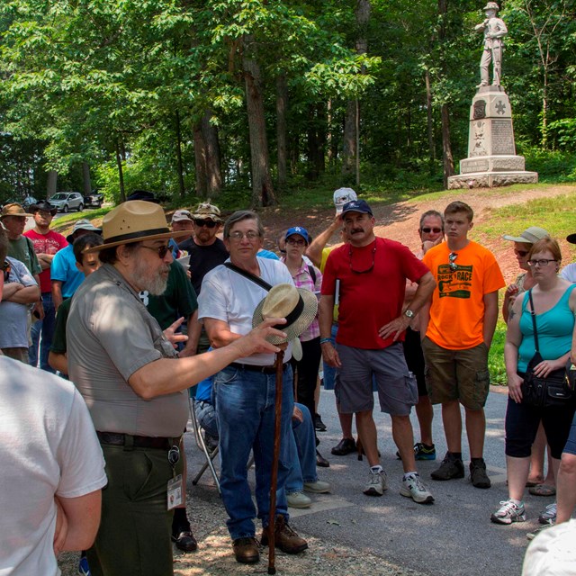 A park ranger presents a program on the battlefield. He is surrounded by dozens of visitors.