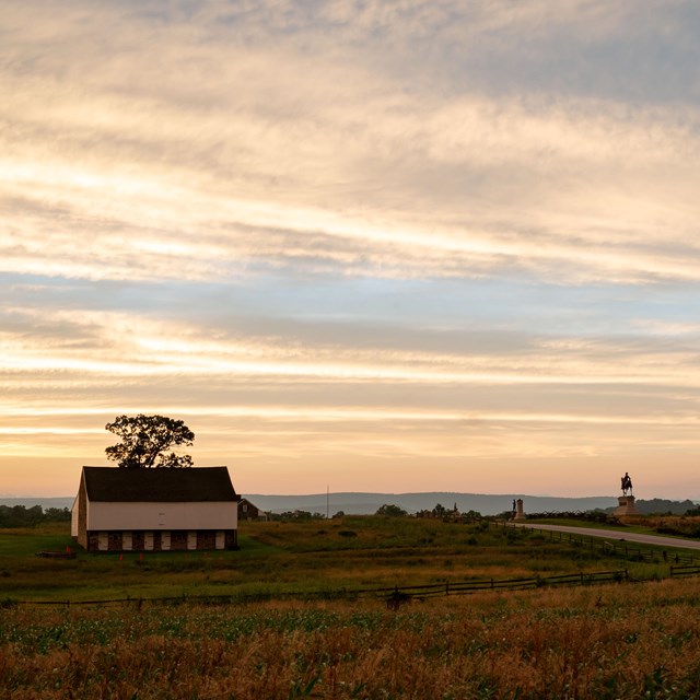 A two story farmhouse next to a road backlit by a sunset.