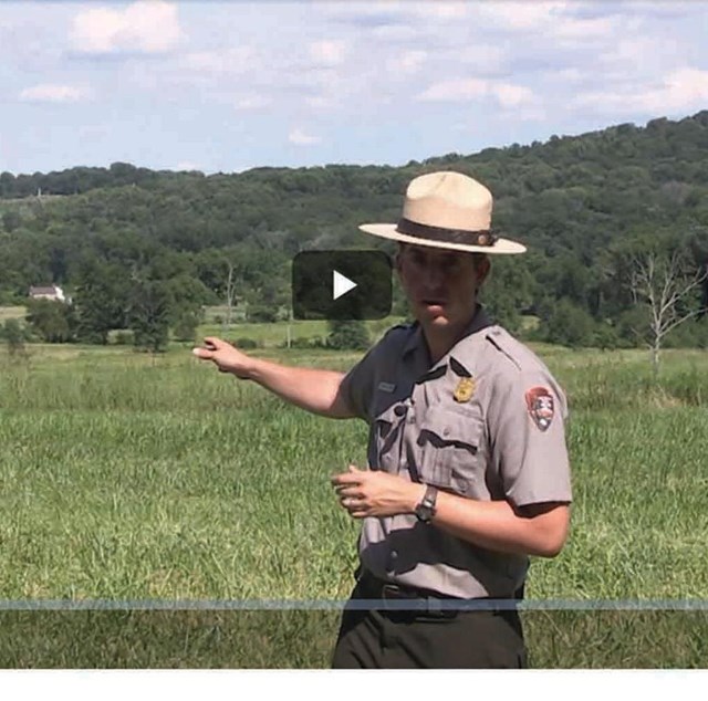 A park ranger points across a green field to two hills in the background. 