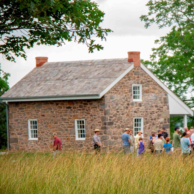 A crowd of visitors stand near a small stone house. Trees are around the house.
