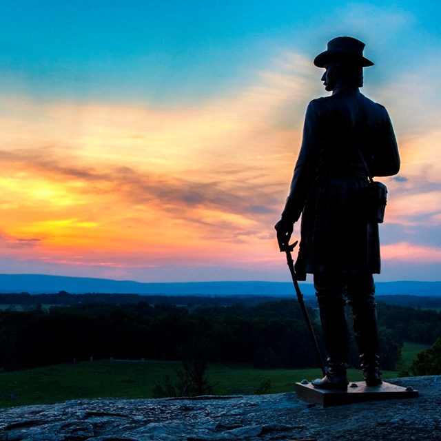 Large statue on boulder overlooking a golden pink sunrise. 