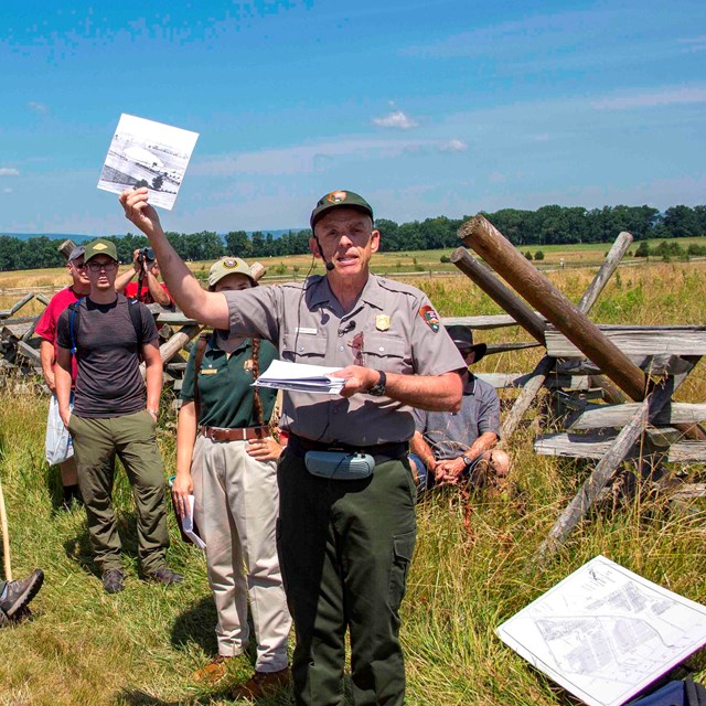 A park ranger shows the crowd a picture during a battle walk in an open field with a wooden fence.