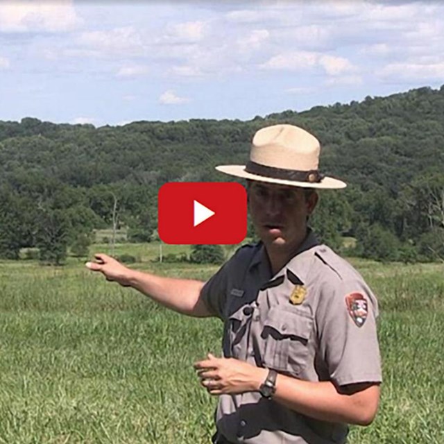 A freeze frame from a video with a ranger in front of a green hill.