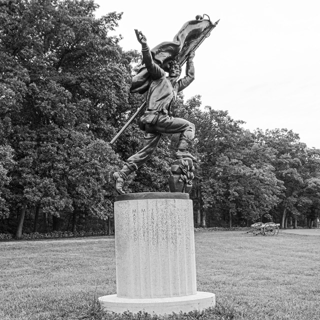A black and white photo of a monument with a man holding a flag with an outstretched right hand. 