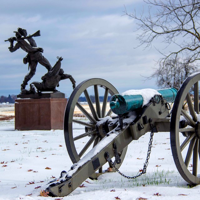 A greenish-turquoise canon appears in front of a large monument that depicts two soldiers.