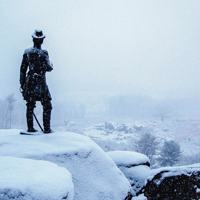 A statue appears on top of a large group of boulders. The landscape is covered in snow.