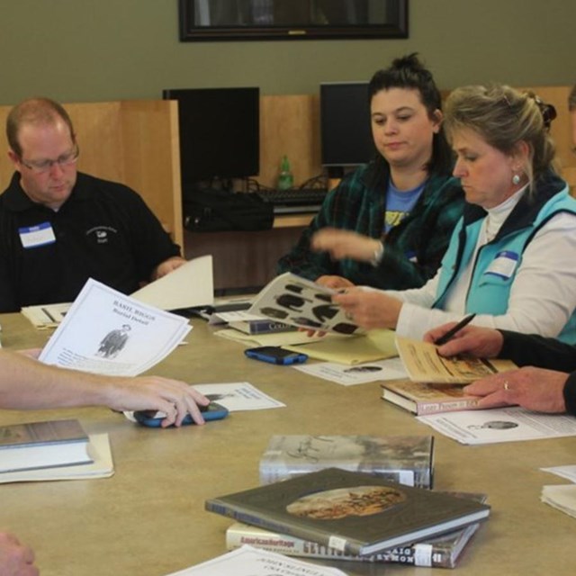 Eight adults gathered around a table with an assortment of books on top.