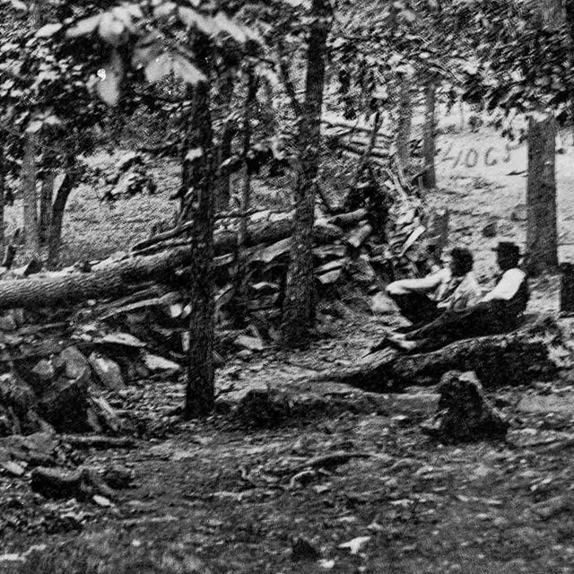 A black and white photo of two men sitting on a rock, among a grove of trees, behind a stone wall.