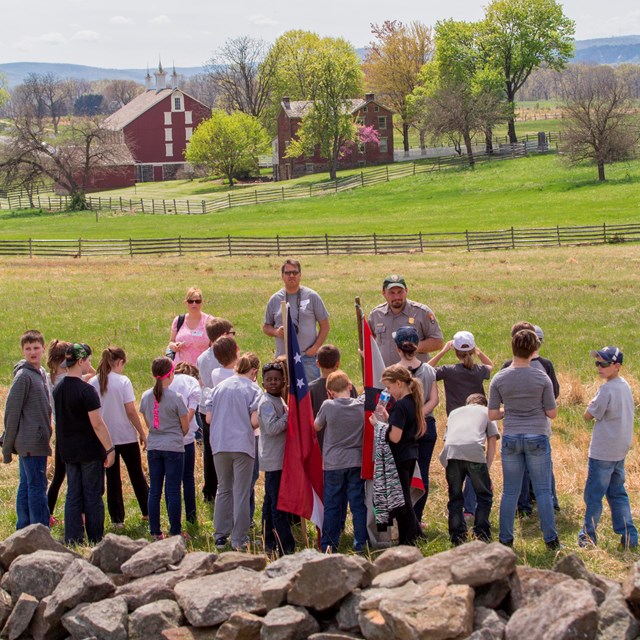 A park ranger leads a group of children on an education program along a stone wall with flags. 