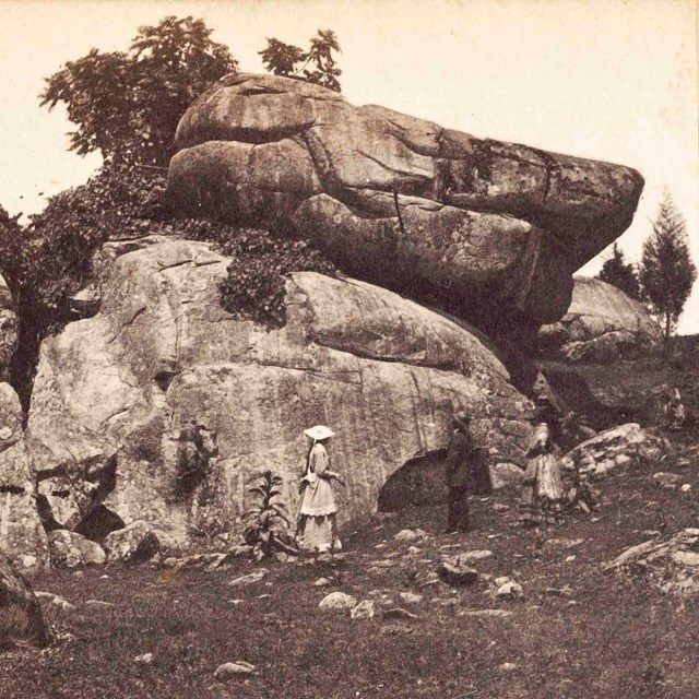 A black and white photo of two women and one man in front of a very large stack of boulders.