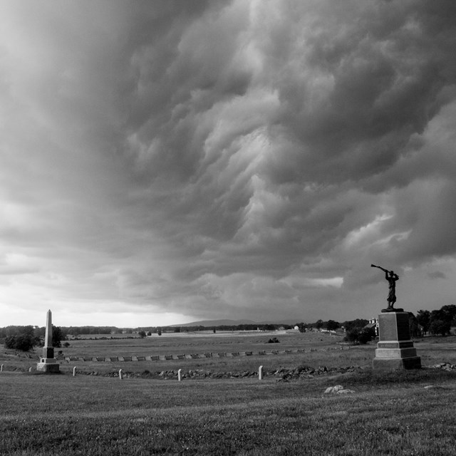 A black and white photo of storm clouds over the battlefield, two monuments are in the foreground.