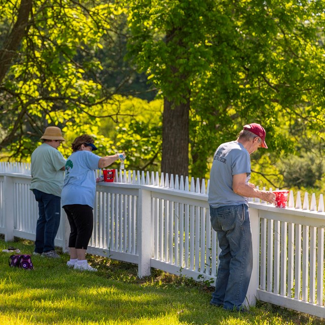 A group of five people repaint a white picket fence. They stand inside the fence in the yard.