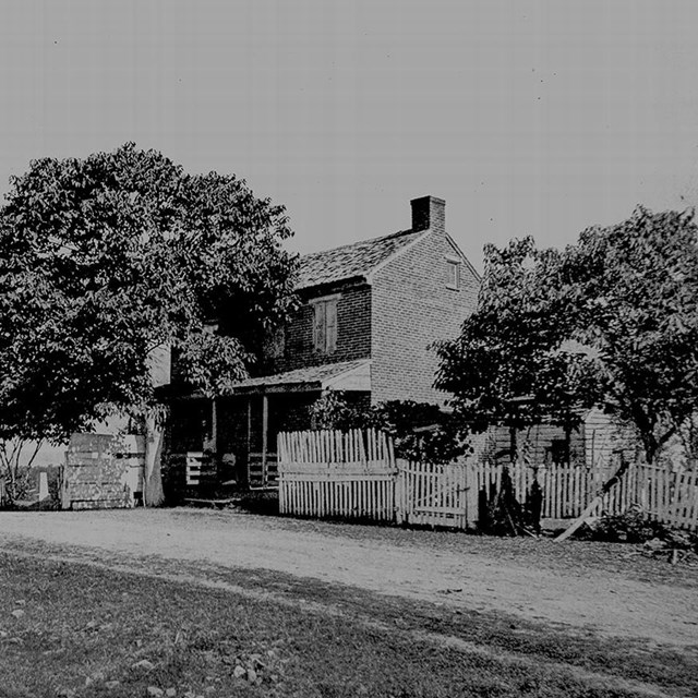 A black and white photo of a brick house sitting next to a dirt road with a white picket fence.