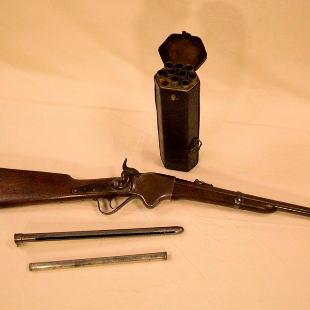 A spencer carbine rifle, ammunition case, and cartridge barrels sit on a table.