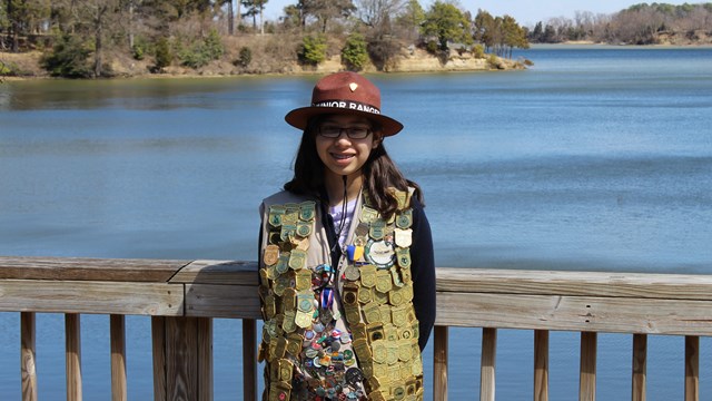 Junior Ranger standing with junior ranger hat and vest filled with badges in front of Popes Creek
