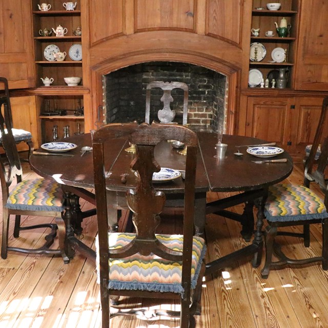 a table and chairs on a wooden floor and wooden walls with a fireplace in the background.