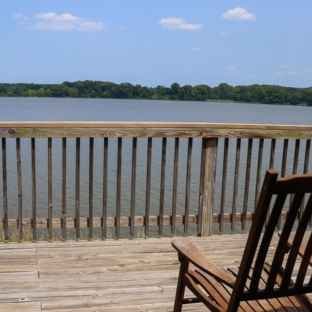 A wooden rocking chair on a wooden deck overlooking beautiful blue water.