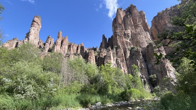 A river runs through a canyon surrounded by lush vegetation, rock spires tower in the background