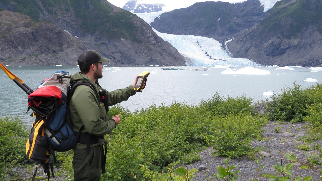 A backcountry park ranger stands on a trail in front of a body of water and glacier collecting data.