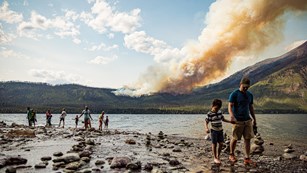 People walk on shore with a wildfire burning across a lake.
