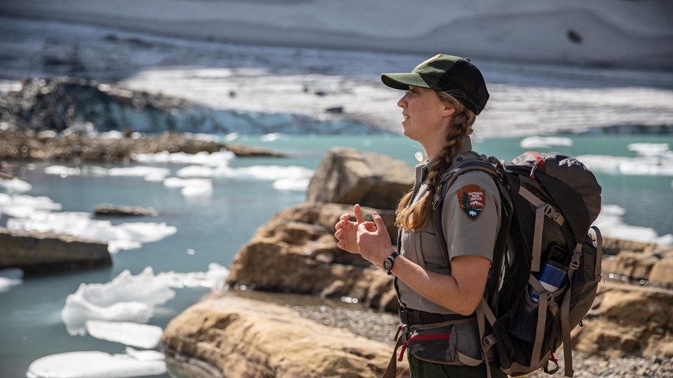 A park ranger stands with their arms out in front of water, rocks, and chunks of ice. 