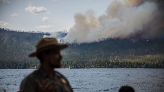 A park ranger stands in front of a lake and a smokey fire. 
