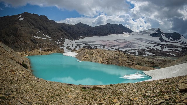 A bright lake surrounded by gravel, snow, and ice in the mountains. 