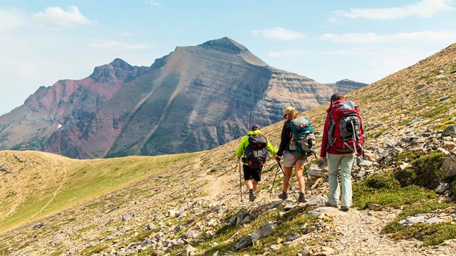 Three people with large backpacks stand on a mountain ridge with mountains in the background.