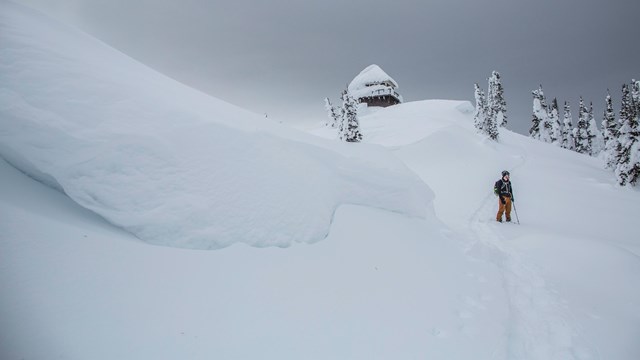 A person stands on a snowy ridgeline before a snow-covered firetower and trees.