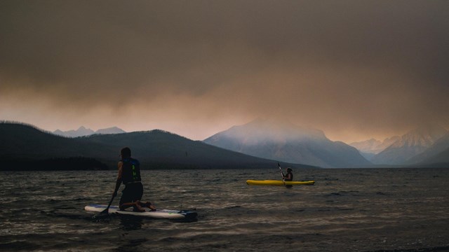 Two people paddle on a dark lake under a thick layer of smoke with mountains in the background.