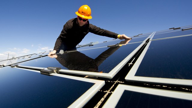 A person in a hardhat reaches out and adjusts one solar panel among many. 