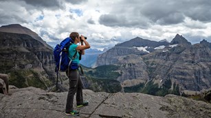 a hiker on a mountaintop looks through binoculars as dark clouds approach