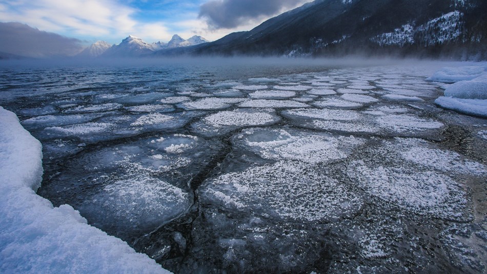 Photo of Lake McDonald with ice in the foreground and snowy mountains in the background.