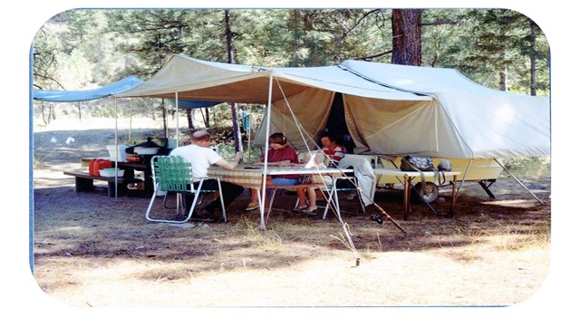 Group of people sit around a picnic table next to an R.V. There are trees around the campsite.