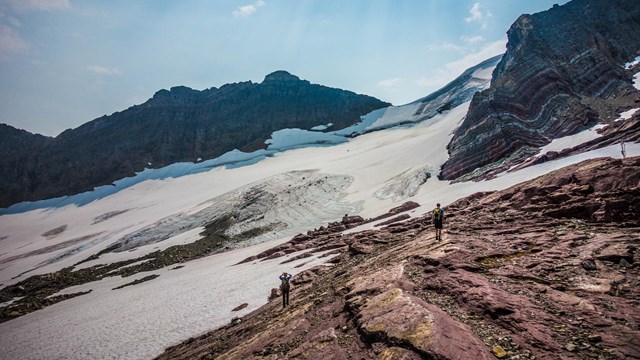People walk over angled rocks toward snow and ice in the mountains. 