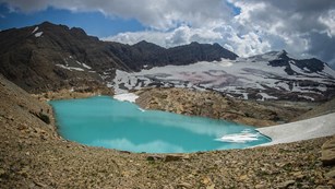 A bright turquoise lake with snow, ice, and mountains in the background. 