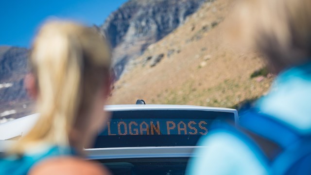 A shuttle bus is parked in front a of a large rocky peak with people in the foreground.