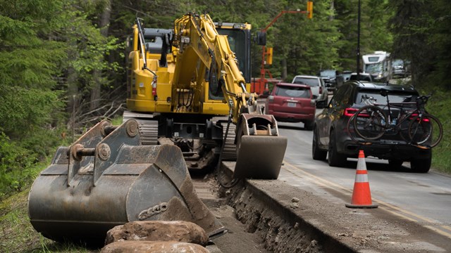 Heavy machinery and bright orange cones along the road. Cars drive by. 