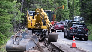 One lane of a road is under construction and occupied by an excavator, while cars travel by.