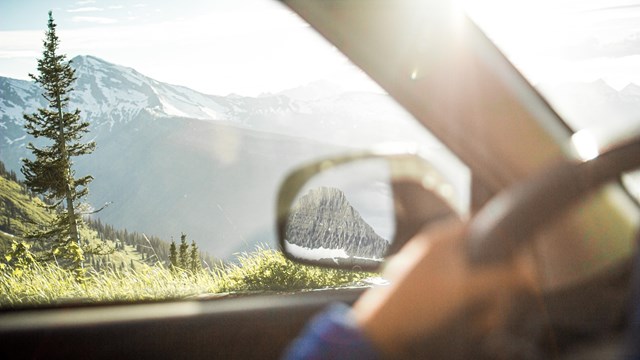 A person's hand on a car's steering wheel with a sun flaring over snowy mountain peaks. 